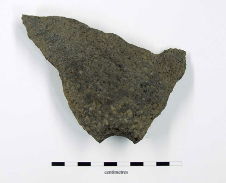 Fragment of lavastone quern with central or handle hole partially intact, some tooling of grinding surface visible. 25mm thick